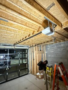 An interior view of a garage under construction featuring an exposed wooden beam ceiling and a partially finished wall with wooden framing and cinder blocks. A modern glass-panelled garage door is visible, with a motorised door opener installed on the ceiling. Various tools, including ladders and a tripod light, are scattered around, along with a box of materials, indicating ongoing work. The garage is well-lit, showcasing the clean concrete floor and clear nighttime view through the glass panels.