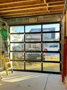 A person in a green shirt stands on a ladder, working on the installation of a modern glass-panelled garage door inside a partially constructed building. Sunlight streams through the transparent sections, highlighting the clean concrete floor and unfinished wooden ceiling beams. Outside, a van branded with "Main Door" and a contact number is parked, with houses and a traffic cone visible in the background.